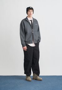 Graue Fleecejacke mit Reißverschluss, hellrosa Hemd, schwarze Hosen mit Taschen und graue Stiefel auf einem blau teppichbedeckten Boden. Schwarze Mütze auf dem Kopf.