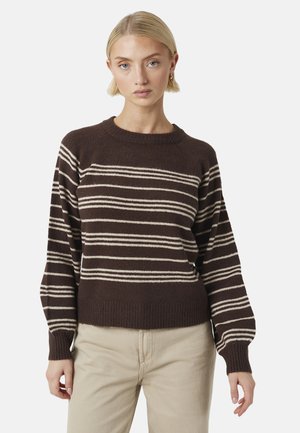 Blondine met een donkerbruine trui met beige horizontale strepen en lichtbeige broek, staand tegen een effen achtergrond.