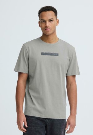 Jauns vīrietis ar īsiem cirtainiem matiem, valkā pelēku T-kreklu ar Blend Chen logotipu un tumšas džinsas, stāvot pret vienkrāsainu gaišu fonu.