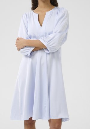 Femme portant une robe ample bleu clair jusqu'aux genoux avec des manches ballon trois quarts et un col en V, bras croisés devant un fond uni.