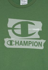 Tissu vert avec le logo et le texte Champion en relief blanc sur un fond ressemblant à du papier déchiré, étiquette de taille visible au col.