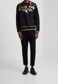 Sweatshirt noir avec motifs décoratifs dorés et texte "VERSACE JEANS" ; présente un col rond, des poignets rayés et un ourlet. Associé à un pantalon et des chaussures noirs.