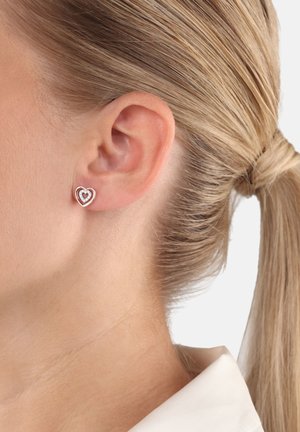 Orecchini a forma di cuore in metallo argentato, con una gemma centrale, contornati da un design a doppio cuore, indossati all'orecchio di un modello.