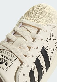 Baskets en cuir blanc avec embout crème texturé, trois bandes noires et accents de couture en zigzag. Dotées de lacets tissés et d'une étiquette de languette de marque.