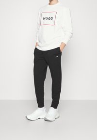Sudadera blanca con el logo negro "HUGO" enmarcado en rojo, combinada con pantalones negros que presentan un logo blanco. El modelo lleva zapatillas blancas.