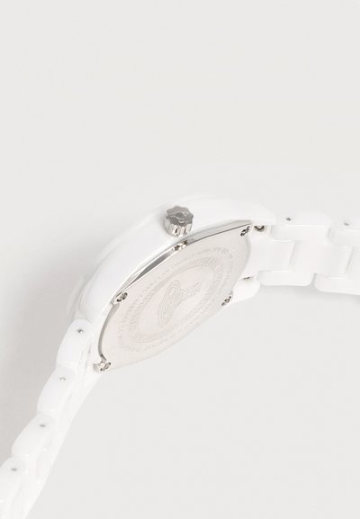 Montre en céramique blanche avec un dos en métal argenté, une couronne texturée et un bracelet à maillons sur un fond clair uni.