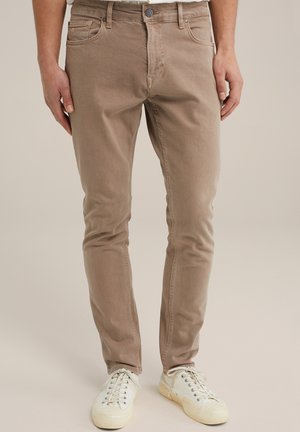 Vaaleanruskeat farkut slim fit -mallilla, joissa on näkyvät ompeleet, etutaskut ja vetoketjukiinnitys. Tyylitetty valkoisilla nauhakenkätrellä.