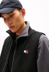 Schwarze Weste mit grauem Kragen, mit durchgehendem Reißverschluss und einem kleinen Tommy Jeans Logo. Der Stoff ist glatt, mit einem weichen, strukturierten Futter.