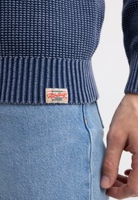 Tamsiai mėlynas megztinis su gipiūro rankovėmis, su Heritage etikete. Sukomplektuotas su šviesiai mėlynomis džinsomis, rodančiomis artimą apžvalgą.