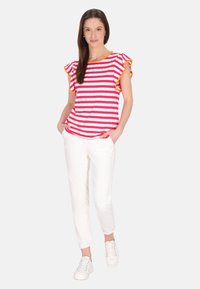 Gestreepte t-shirt in roze en wit met ruches aan de mouwen en oranje accenten, gecombineerd met witte broek en witte sneakers.