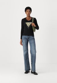 Svart langermet skjorte med et trekantet design av paljetter, kombinert med blå flared jeans og svarte sko. Holder en grønn veske.
