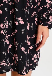 Robe en tissu noir ornée de motifs floraux roses et gris, avec une coupe ample et un détail de manchette boutonnée sur la manche.