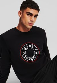 Felpa nera con collo a coste; presenta un logo circolare bianco e rosso, ben visibile sul petto, in tessuto di cotone morbido.