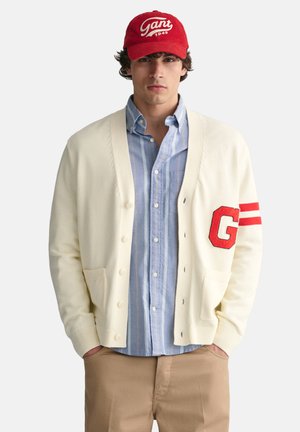 Gilet crème avec la lettre rouge "G" et des rayures, porté sur une chemise à rayures bleues. Associé à une casquette rouge avec un logo blanc. Look décontracté.