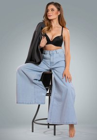 Reggiseno imbottito nero, pantaloni ampi in denim blu chiaro oversize e un blazer grigio appoggiato sulla spalla, seduta su uno sgabello.