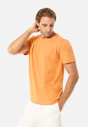 Homme aux cheveux châtain clair portant une chemise orange à manches courtes et un pantalon blanc, une main derrière la nuque et l'autre dans la poche.