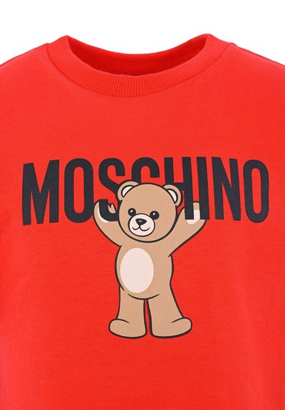 Czerwona bawełniana koszulka z kartonowym misiem z uniesionymi ramionami oraz napisem "MOSCHINO" w grubych czarnych literach nad misiem.
