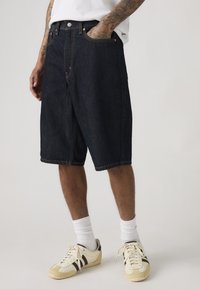 Άνδρας με σκούρες μπλε τζιν βερμούδες μέχρι το γόνατο, λευκές κάλτσες μέχρι τον αστράγαλο, μπεζ και καφέ αθλητικά παπούτσια, με τατουάζ στα χέρια και το ένα χέρι στην τσέπη.