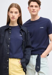 Νεαρή γυναίκα και άνδρας φοράνε μπλε μπλούζες ARKK. Η γυναίκα φοράει τζιν πουκάμισο και κίτρινες παντελόνες, ο άνδρας έχει ανοιχτόχρωμα τζιν με ριγέ γραβάτα στην τσέπη.