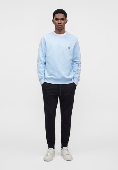 Homme debout, les mains dans les poches, portant un sweat-shirt bleu clair, un pantalon de jogging noir et des baskets blanches sur un fond uni.