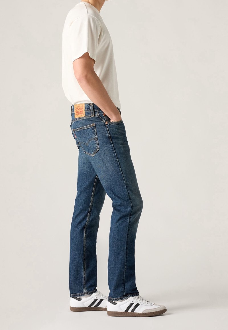 Personne portant un jean droit bleu, un t-shirt blanc et des baskets blanches avec des bandes noires, debout de profil.