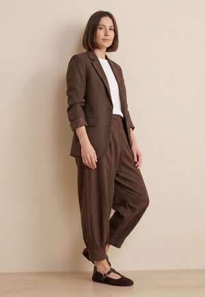 Woman standing against beige wall wearing brown checked blazer, white shirt, loose brown trousers, and dark brown Mary Jane shoes.