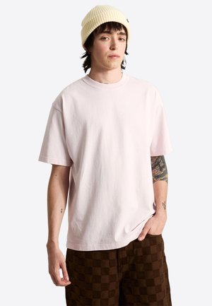Jeune personne portant un t-shirt oversize rose clair, un bonnet en tricot beige et un pantalon marron à carreaux, avec des tatouages visibles sur les bras.