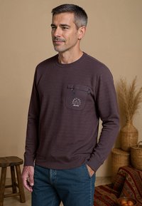 Donkerpaarse lange mouwen shirt met een gestructureerde afwerking, voorzien van een borstzak en grafische details. Gecombineerd met blauwe spijkerbroek. Achtergrond is neutraal.