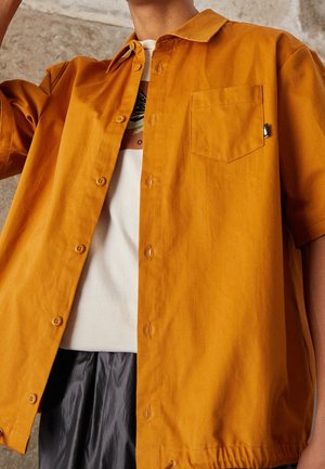 Homme portant une chemise ouverte jaune moutarde à manches courtes sur un t-shirt blanc imprimé et un pantalon sombre devant un mur texturé.