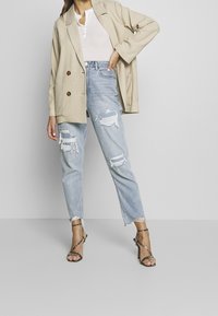 Beige oversized blazer with two buttons, white textured top, and light blue distressed jeans featuring frayed hems and ripped patches. Black strappy heels.