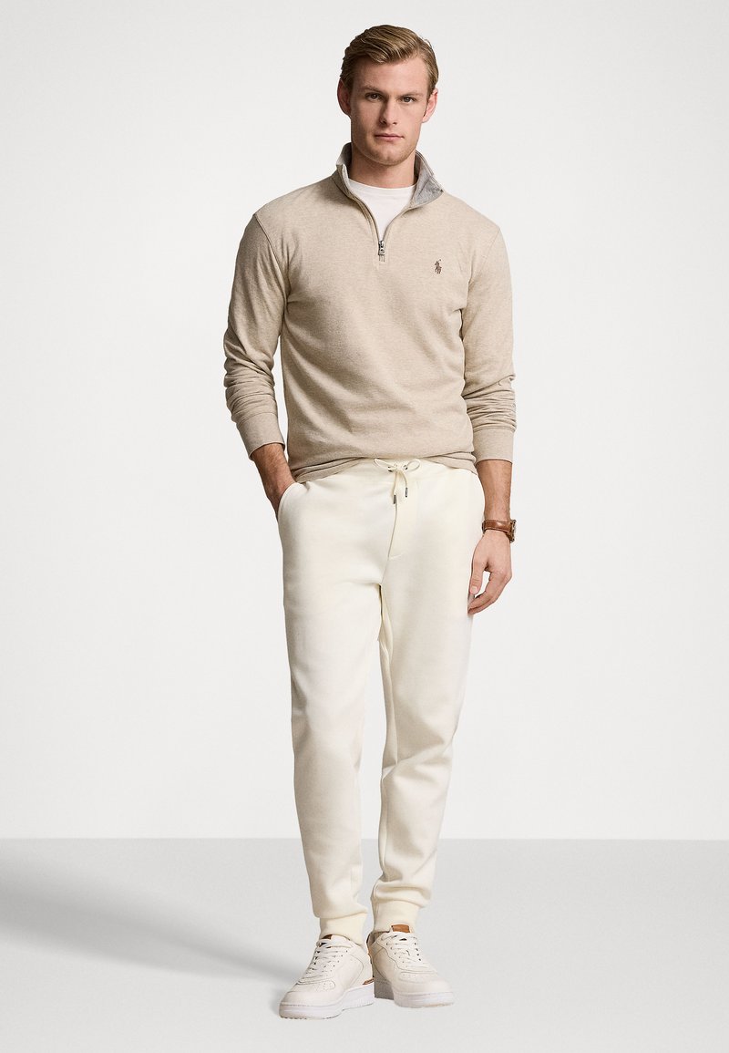 Maglione beige con zip e colletto grigio chiaro, abbinato a pantaloni felpati bianchi a gamba affusolata e sneakers bianche. Design semplice, senza motivi o dettagli.