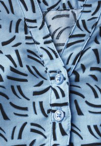 Tessuto blu chiaro con motivi astratti neri, con una chiusura a bottoni e una texture morbida. Include un leggero dettaglio a bottone.