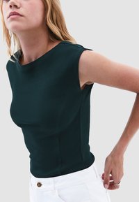 Top senza maniche di un verde scuro a costine con scollo rotondo e design aderente, abbinato a pantaloni bianchi con un dettaglio in bottoni dorati.