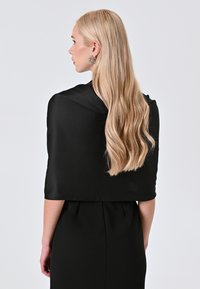 Zwarte satijnen sjaal valt over de schouders, met een gladde textuur en wijd, cropped ontwerp. Lange, golvende blonde haren vloeien langs de rug naar beneden.