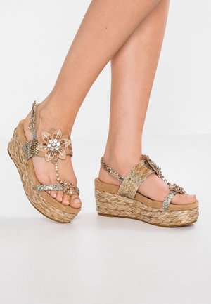 Benen met gouden sleehak sandalen met gevlochten plateauzolen en juweelachtige bloemdecoraties op een lichte huidskleur.