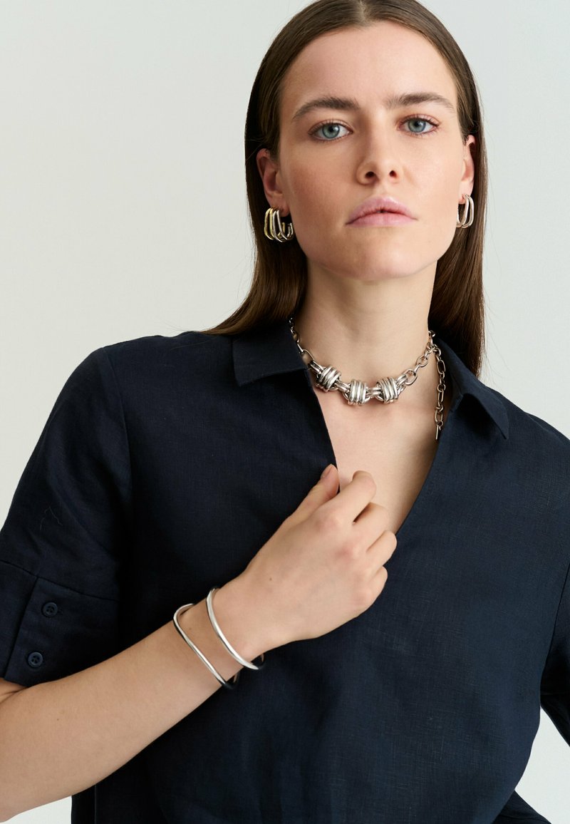 Femme aux cheveux bruns et lisses portant des boucles d'oreilles créoles en argent, un collier chaîne argentée épaisse, des bracelets en argent et une chemise sombre à col.