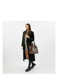 Hnědá kožená taška přes rameno se strukturovaným designem, dvěma uchy a jemnou texturou, doplněná dlouhým černým kabátem přes šedé šaty a černé boty.