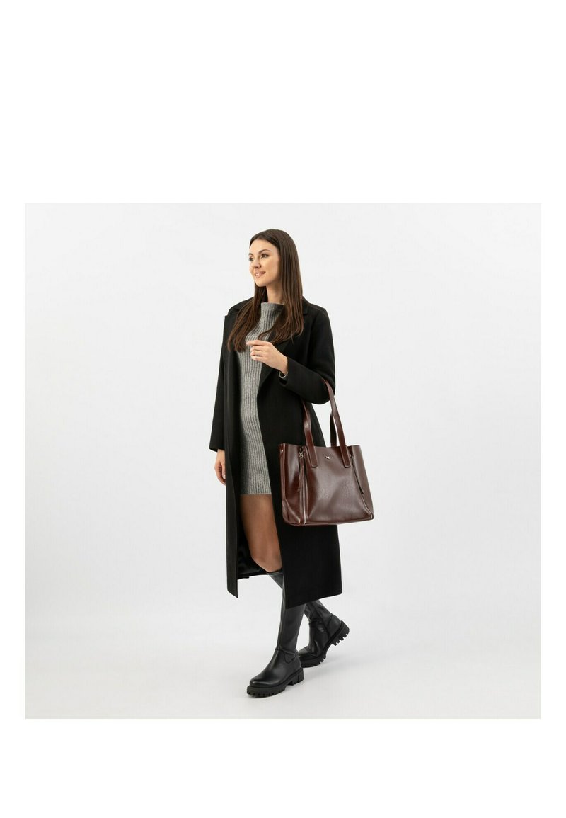 Hnědá kožená taška přes rameno se strukturovaným designem, dvěma uchy a jemnou texturou, doplněná dlouhým černým kabátem přes šedé šaty a černé boty.