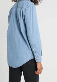 Ljust blå denimskjorta med långa ärmar, knäppte manschetter, en rundad fåll och detaljseams vid ryggen. Bärs med svarta byxor.