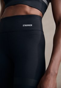 Detailní záběr osoby, která má na sobě černé legíny s vysokým pasem s nápisem "STRONGER" vytištěným na pásku a odpovídající černý sportovní top.