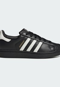 Czarny skórzany sneakers z białym futrzanym akcentem, trzema białymi paskami, gumową podeszwą i czarnymi sznurowadłami. Widoczne złote logo "SUPERSTAR".