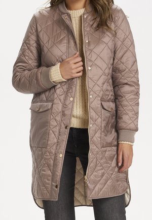 Vrouw draagt een lange, doorgestikte taupekleurige jas met drukknopen en rits, een beige gebreide trui en een donkere spijkerbroek, terwijl ze de revers van de jas vasthoudt.