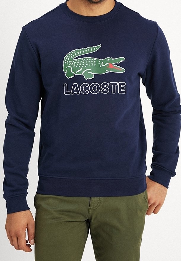Homme portant un sweat-shirt bleu marine avec un grand logo crocodile vert et le texte "LACOSTE", associé à un pantalon vert olive.