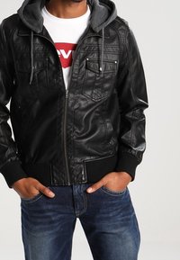 Svart läderjacka med grå huva, två bröstfickor, dragkedjestängning, ribbade mudd och en lös passform, bärs över en vit t-shirt.