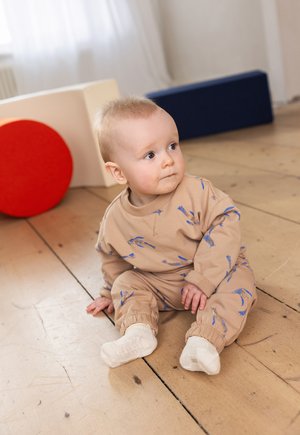 Hellbraunes Kleinkinderoutfit mit blauen Vogelmustern, langen Ärmeln und Bündchen an den Knöcheln. Weicher Stoff, getragen mit weißen Socken auf einem Holzboden.