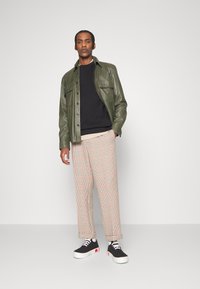 Giacca camicia in pelle verde oliva sopra un maglione nero, abbinata a pantaloni a quadri beige e scarpe da ginnastica nere con suola bianca e dettagli rossi.