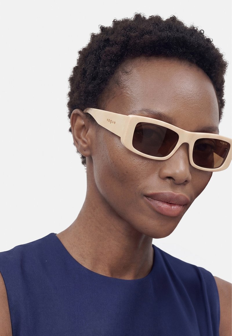 Femme aux cheveux courts et bouclés portant des lunettes de soleil rectangulaires beiges et un haut bleu foncé sans manches sur fond uni.