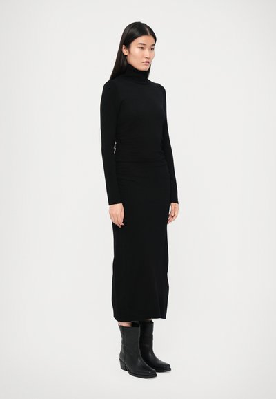 Fekete garbó hosszú ujjakkal, testhez simuló szabású, hosszú fekete szoknyával és bokáig érő fekete csizmával párosítva, így létrehozva egy egységes sziluettet.