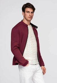 Burgunderfarbene Zip-Jacke mit geripptem Muster und schwarzen Akzenten über einem cremefarbenen Kabelstrickpullover, kombiniert mit weißen Hosen.