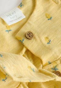 Żółty dziecięcy garment wykonany z teksturowanej tkaniny, z kwiatowym wzorem, w którym znajdują się małe cytryny i niebieskie akcenty. Zawiera detal z drewnianym guzikiem.